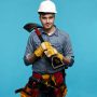 Farming, agriculture and gardening concept. Square picture of positive young male gardener going to dig bed to plant vegetables in garden, carrying shovel on his shoulder, wearing helmet and gloves
